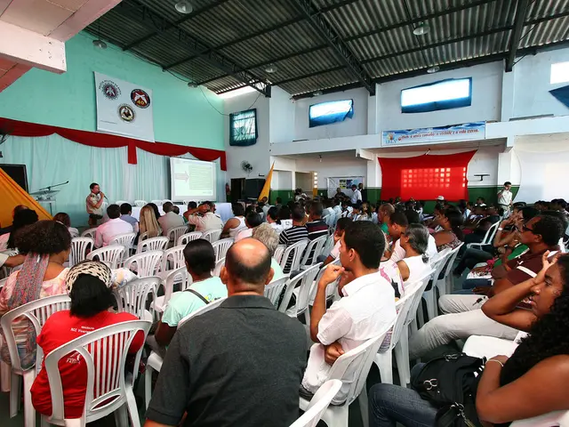 In this picture there are group of people there are sitting on the chairs there is a stage, one...
